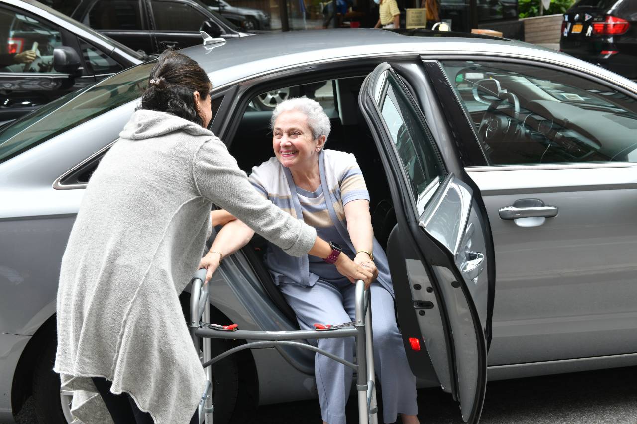 Una mujer con cabello corto canoso recibe ayuda para salir de un vehículo, con un andador.