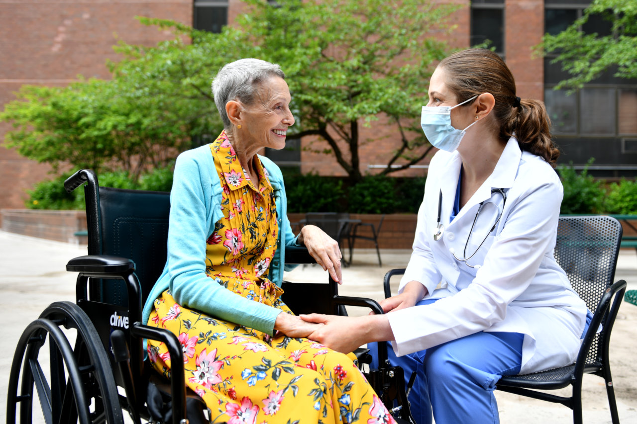 Un residente en una silla de ruedas habla con un enfermero con una máscara en un patio.