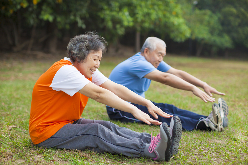 An older woman and man sit in the grass outdoors, legs outstretched, reaching their toes