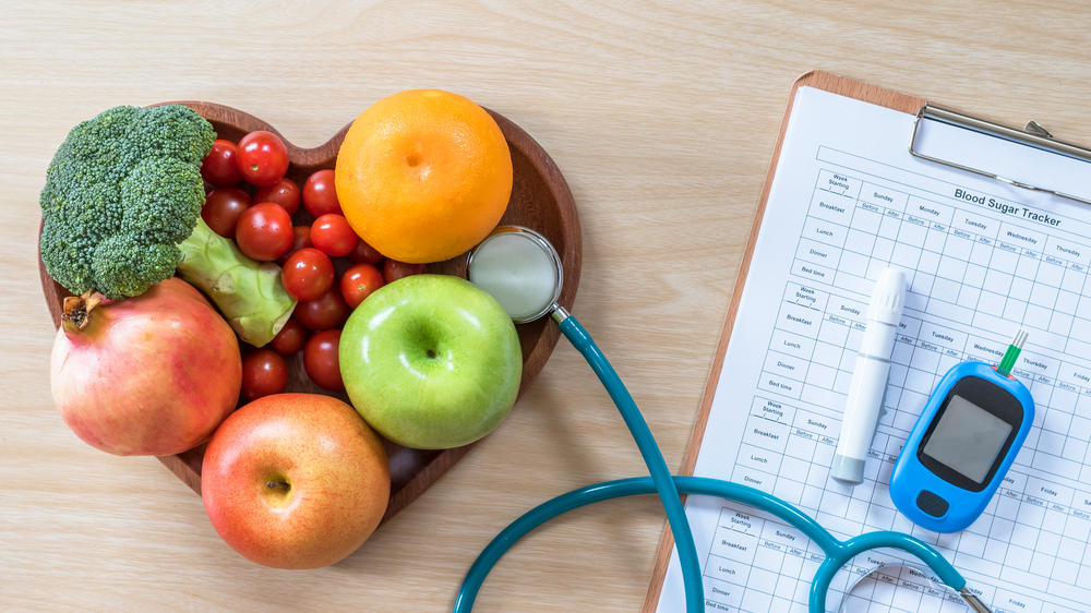 Fruits and vegetables fill a wooden, heart-shaped bowl next to a diet log and blood sugar tracking devices